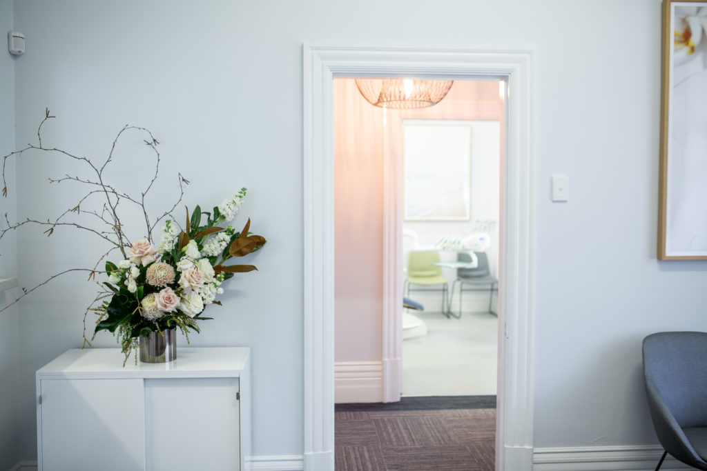 Interior photo of Ailin Teo Dental Surgery. It shows a beautiful arrangement of pink and white flowers in a vase next to a doorway which reveals the pale pink walls of a room across the hallway. Used to emphasise aesthetics.