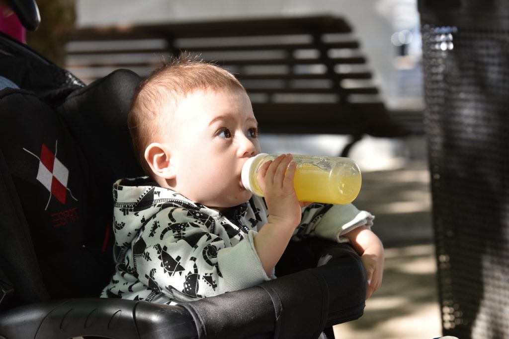A baby sits in a pram holding and drinking from a bottle filled with fruit juice